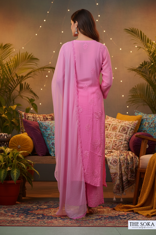 Woman in a pink traditional outfit standing in a decorated living room with plants and colorful decor.