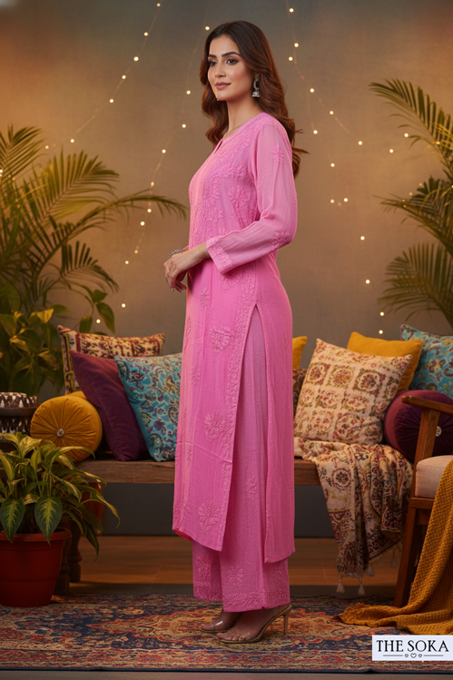 Woman in a pink traditional outfit standing in a decorated room with plants and colorful cushions.