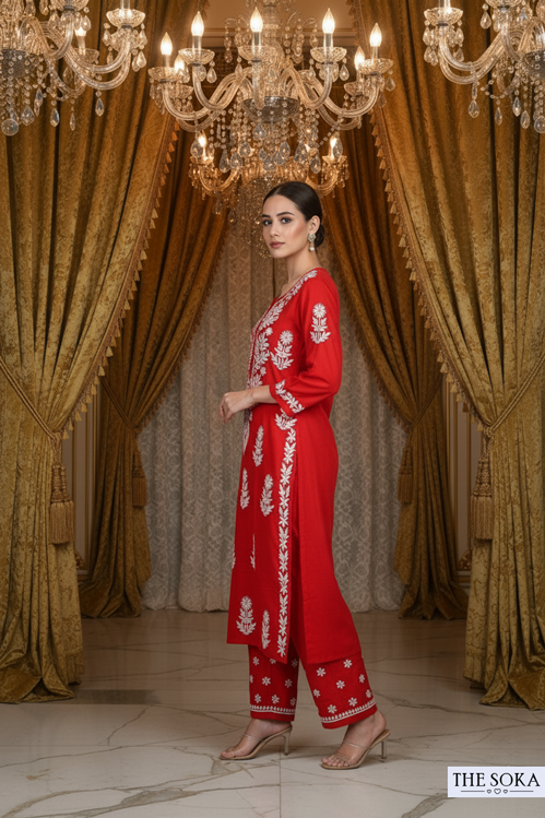 Woman in a red traditional outfit with white embroidery standing in a luxurious room with chandeliers and gold curtains.
