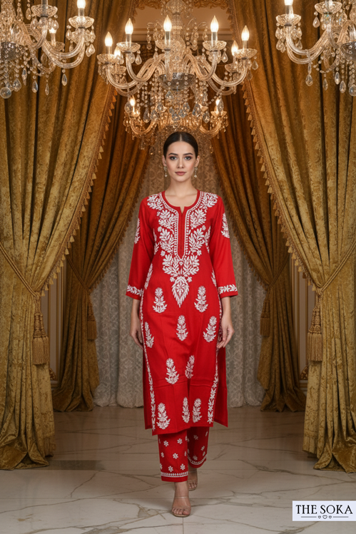 Woman in a red embroidered outfit standing in front of ornate chandeliers and gold curtains.
