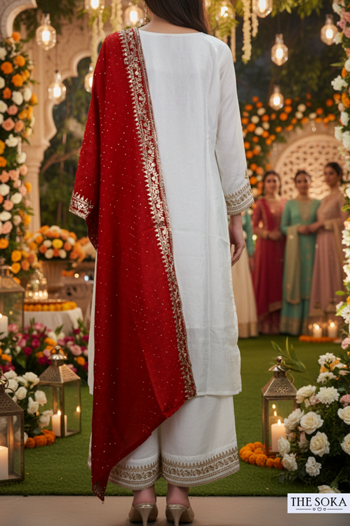 Person wearing a white outfit with a red dupatta in a decorated setting with flowers and lights.