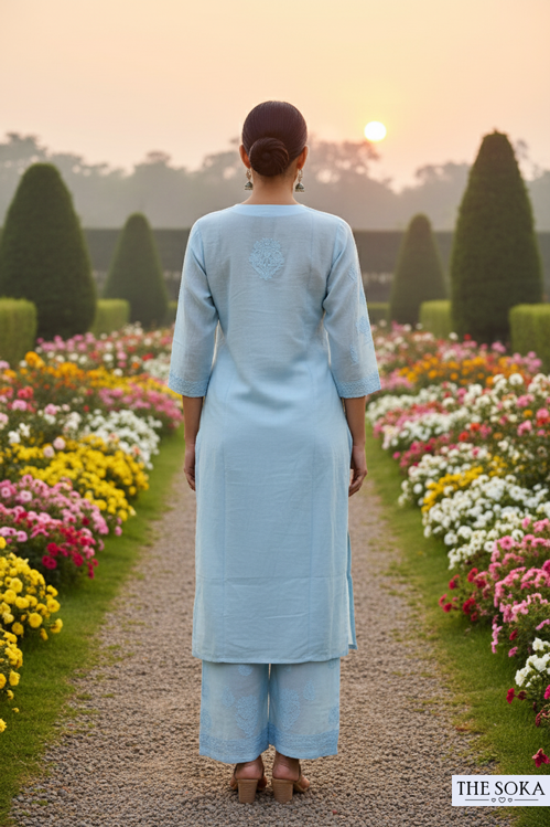 Person in a light blue outfit walking away from the camera through a garden with colorful flowers at sunset.