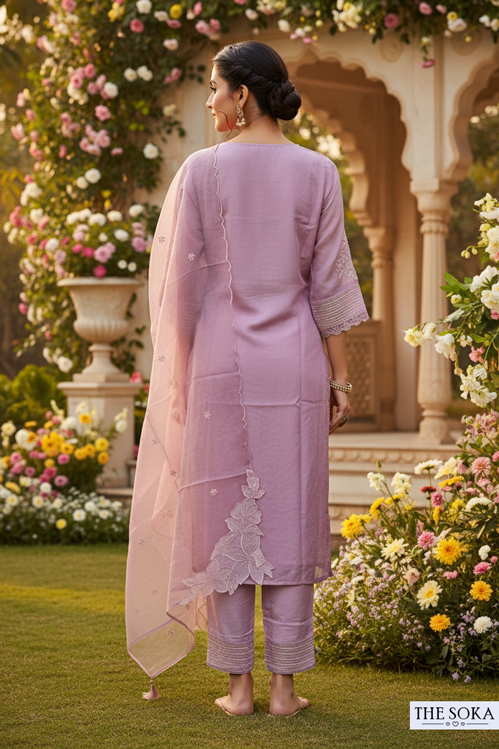 Woman in a lavender traditional outfit standing in a garden with flowers and architectural elements.