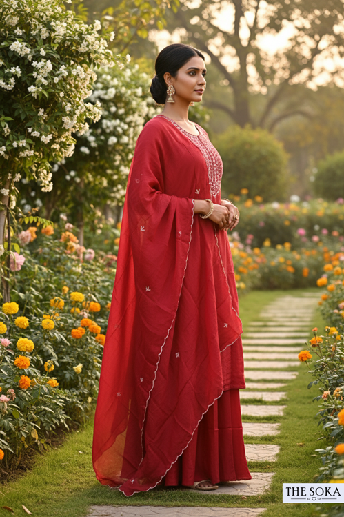 Woman in a red saree standing in a garden with flowers and greenery.